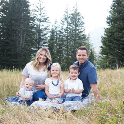 Family of five posing on a blanket in a grassy meadow, smiling at the camera with trees in the background.